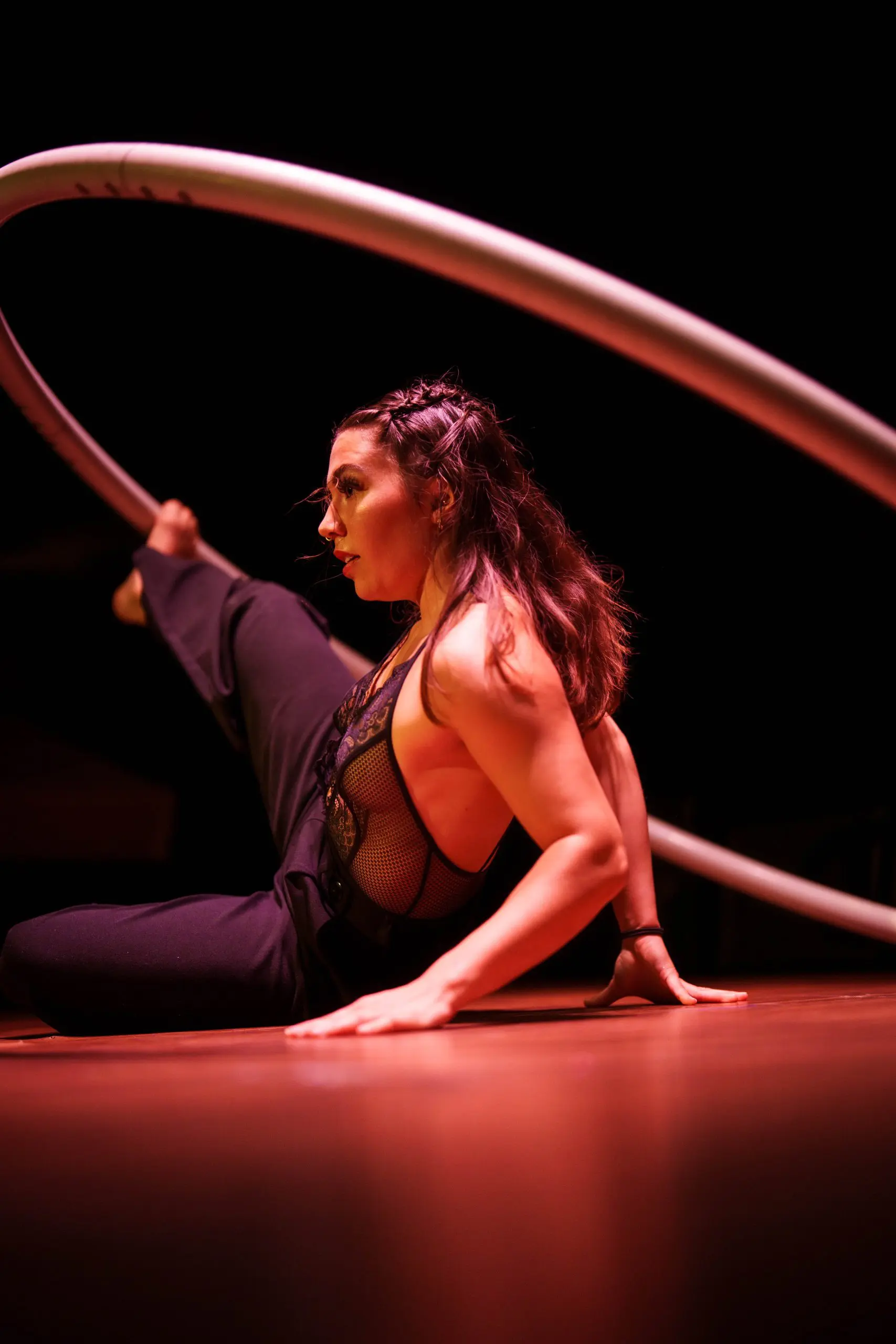 Léonie Salcines, vêtue d'une tenue en maille noire, prend une pose de contorsion flexible sur scène, tenant un grand cerceau métallique avec une jambe levée en hauteur sous un éclairage dramatique.