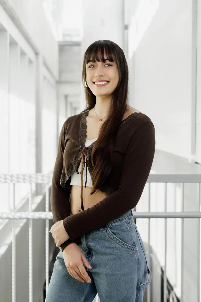 Une jeune femme aux longs cheveux bruns, vêtue d'un gilet brun par-dessus un haut blanc et d'un jean bleu, sourit dans un couloir intérieur moderne et lumineux.