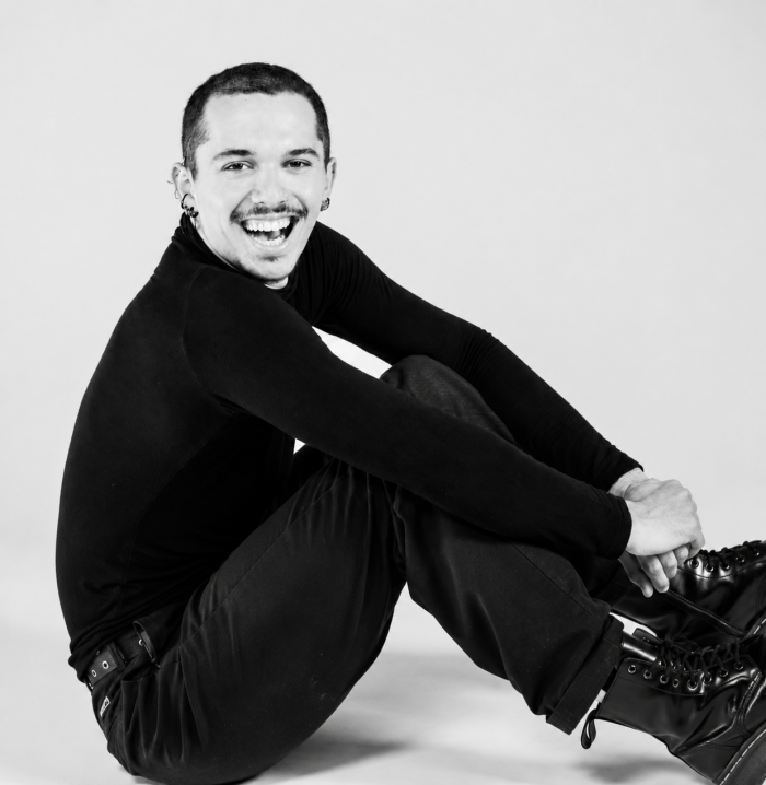 Mathis Machuré, aux cheveux courts et à la moustache, tout de noir vêtu, est assis par terre, le sourire aux lèvres, les bras posés sur les genoux, chaussé de bottines à lacets. Cette photo en noir et blanc immortalise un moment à la fois décontracté et élégant.