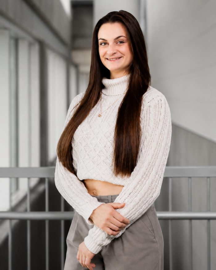 Téa Tkach, une femme aux longs cheveux châtains, vêtue d'un pull à col roulé en maille couleur crème et d'un pantalon gris, se tient à l'intérieur près d'une balustrade, les bras croisés, un sourire aux lèvres. En arrière-plan, on aperçoit de grandes fenêtres et un couloir moderne.