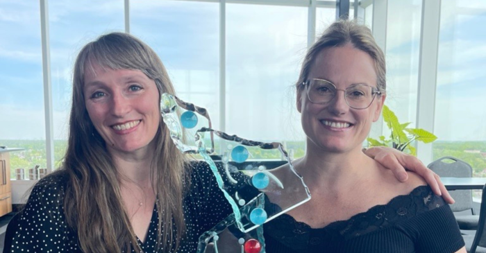 Deux femmes souriantes sont assises côte à côte à l'intérieur, près de grandes fenêtres, tenant une sculpture en verre composée de sphères bleues et rouges. Elles célèbrent le fait que l'École nationale de cirque ait remporté le Prix de l'innovation 2026 de l'ACPQ pour le projet « Rig'Humaine ».