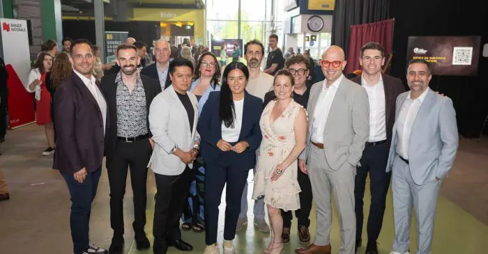 Un groupe de douze personnes, élégamment vêtues en tenue professionnelle ou semi-formelle, sourient pour une photo lors de la Soirée-bénéfice 2026, au cours de laquelle 150 000 dollars ont été récoltés pour soutenir la prochaine génération à l’École nationale de cirque. Des affiches et une enseigne de bistrot ajoutent du charme à cet espace événementiel moderne.