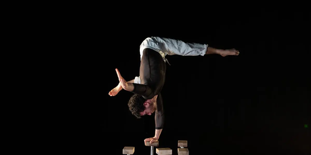 Un acrobate masculin est en équilibre sur ses mains au sommet de quatre barres parallèles, les jambes écartées en grand écart, sur un fond sombre.