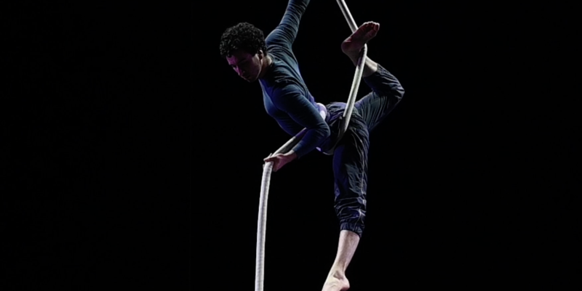 Une personne exécute un numéro d'acrobatie aérienne, suspendue et en équilibre sur une corde blanche, sur fond sombre.