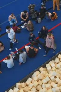 Un groupe de personnes est assis ou debout sur un tapis bleu ; certaines discutent, d’autres se reposent, dans le cadre des auditions de l’ÉNC. Des blocs de mousse occupent le bord inférieur, et une ligne rouge traverse la scène.
