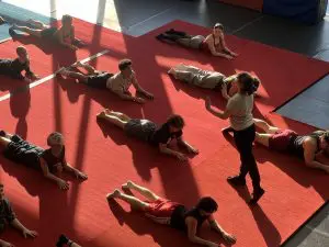 Un groupe de personnes allongées sur le ventre sur des tapis de gym rouges, tandis qu’une personne se tient debout et fait des gestes, sans doute pour donner des instructions ou des conseils — peut-être lors des auditions de l’ÉNC. La lumière du soleil pénètre dans la pièce, projetant des ombres sur la scène.