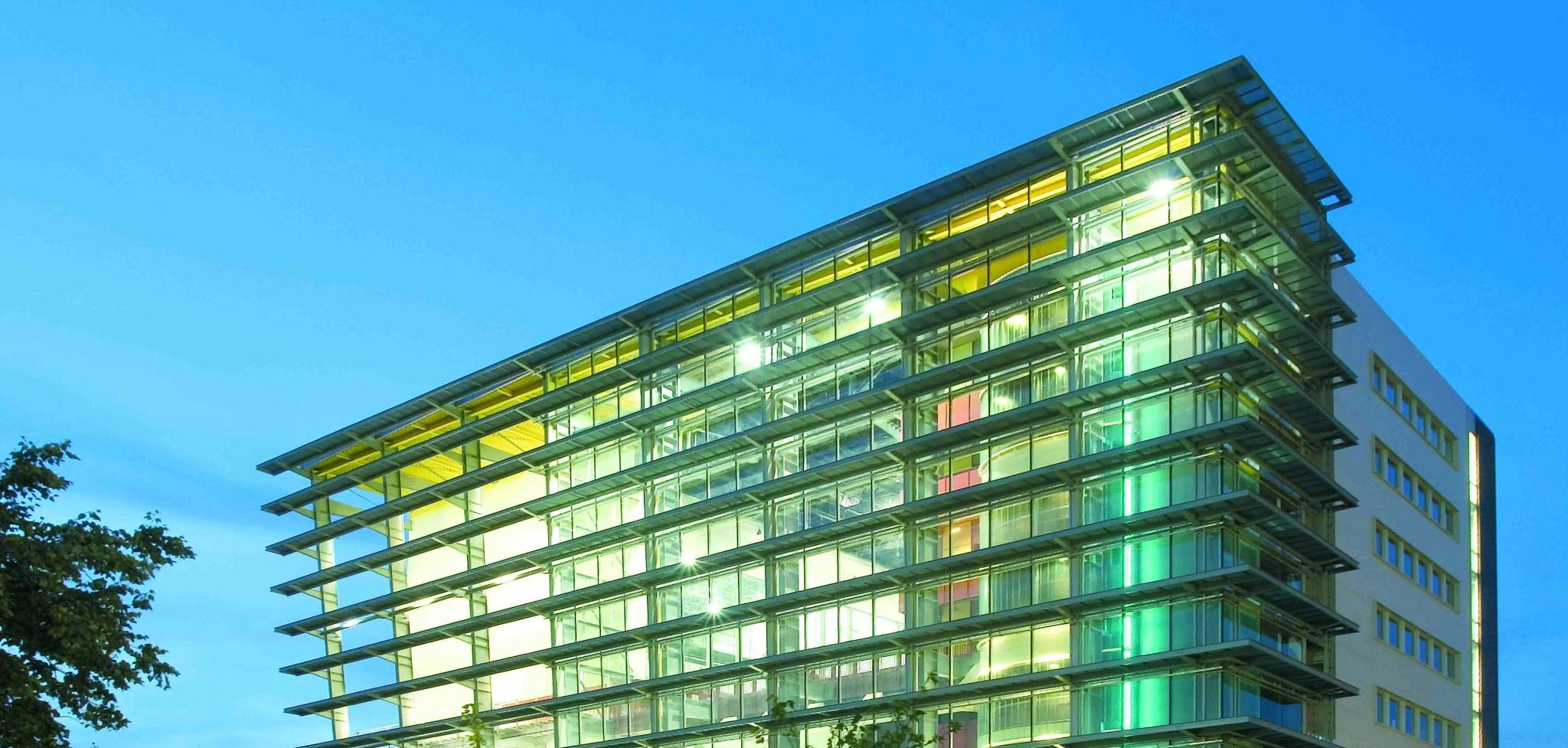 Un immeuble de bureaux moderne en verre, éclairé de l'intérieur au crépuscule, se dresse au milieu d'une pelouse verte et de jeunes arbres sous un ciel bleu clair en soirée - une scène inspirante pour ceux qui sont prêts à rejoindre l'ENC.
