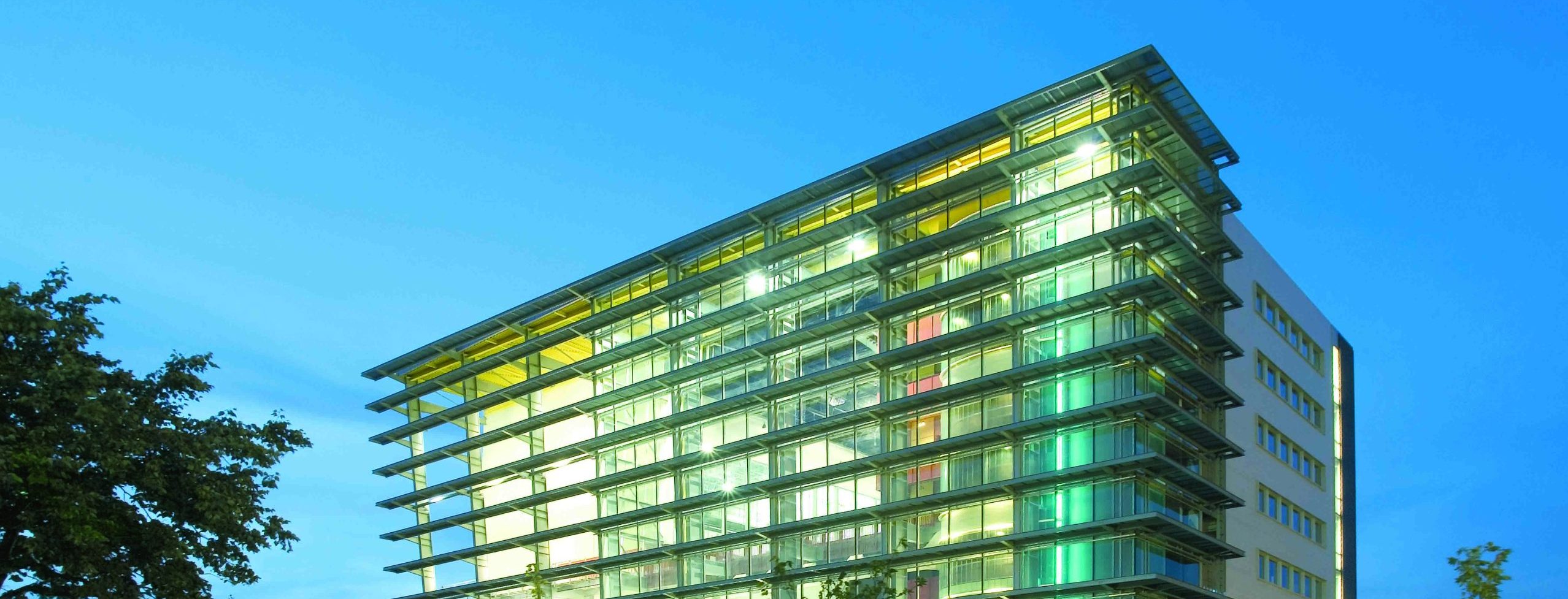 Un immeuble de bureaux moderne en verre, éclairé de l'intérieur au crépuscule, se dresse au milieu d'une pelouse verte et de jeunes arbres sous un ciel bleu clair en soirée - une scène inspirante pour ceux qui sont prêts à rejoindre l'ENC.