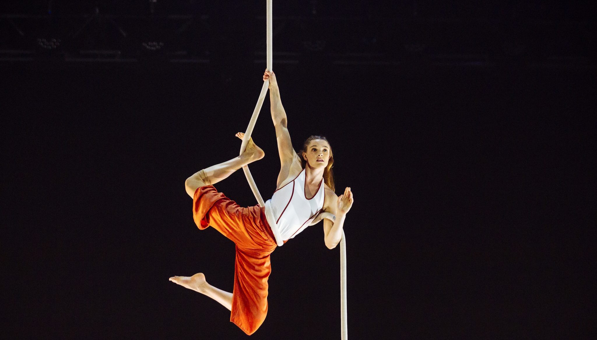 Un artiste aérien en pantalon orange et chemise blanche se suspend gracieusement à une corde blanche sur fond sombre, vous invitant à découvrir l'École en équilibre avec une jambe crochetée et un bras tendu vers le haut.