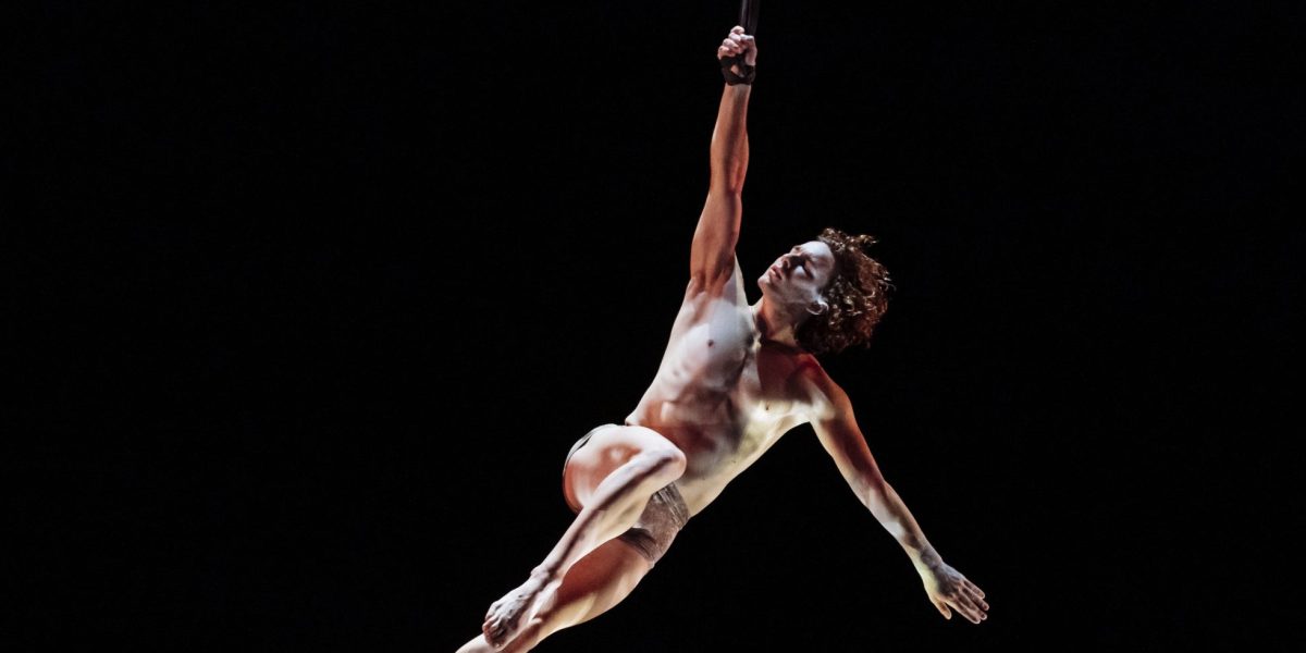 L'artiste aérien Agustin Rodriguez Beltran est suspendu dans les airs, se tenant à une corde d'une main, le corps allongé et illuminé sur un fond sombre.