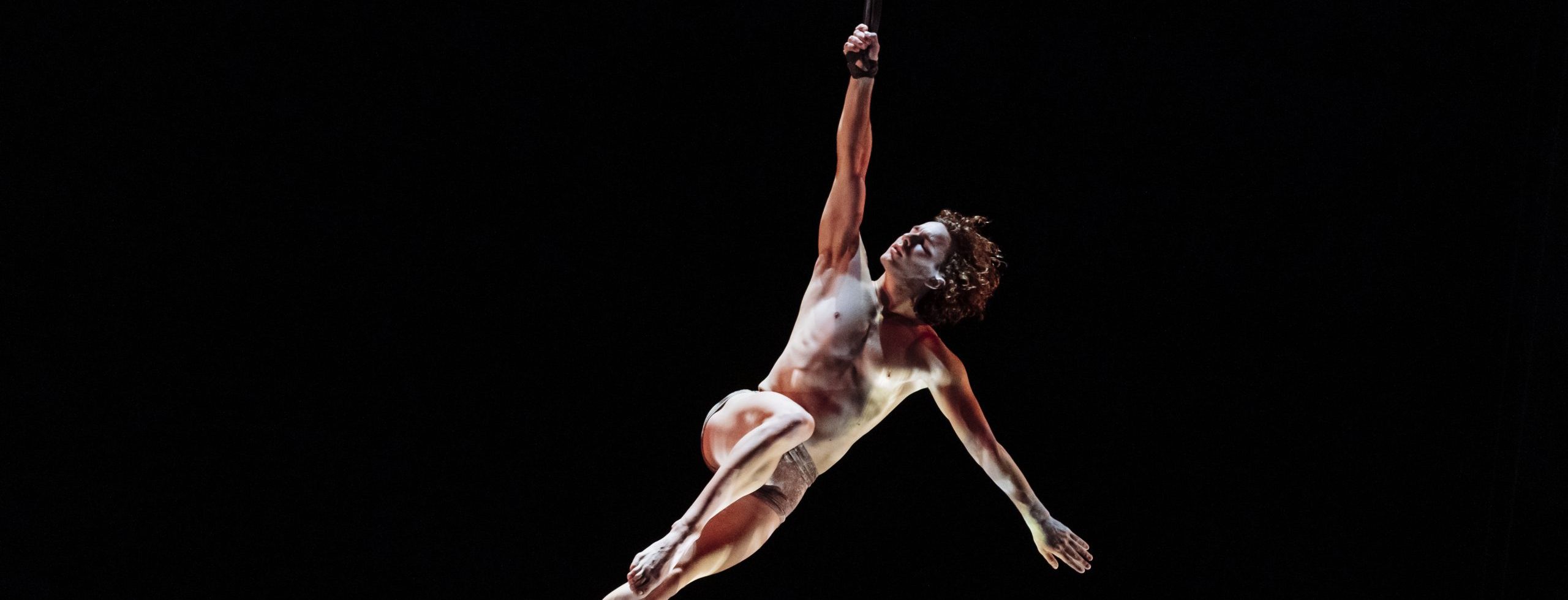 L'artiste aérien Agustin Rodriguez Beltran est suspendu dans les airs, se tenant à une corde d'une main, le corps allongé et illuminé sur un fond sombre.