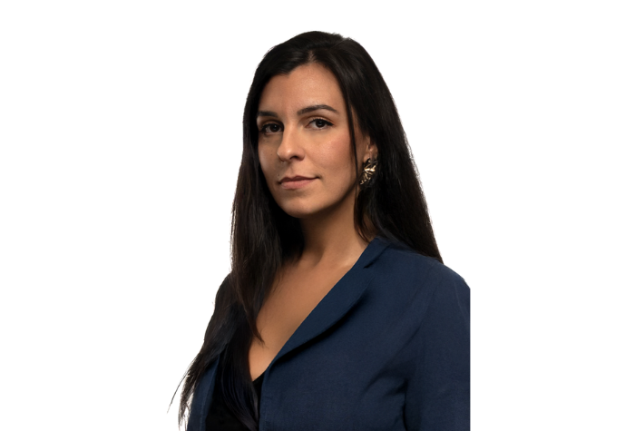 Alicia Bremond, aux longs cheveux noirs, vêtue d'un blazer bleu foncé et portant des boucles d'oreilles en or, se tient debout sur un fond blanc uni, regardant l'appareil photo avec assurance.