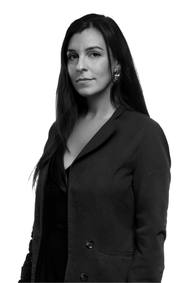 Portrait en noir et blanc d'Alicia Bremond, une femme aux longs cheveux noirs, portant un blazer sombre sur un haut, debout et regardant l'appareil photo avec une expression neutre sur un fond blanc uni.