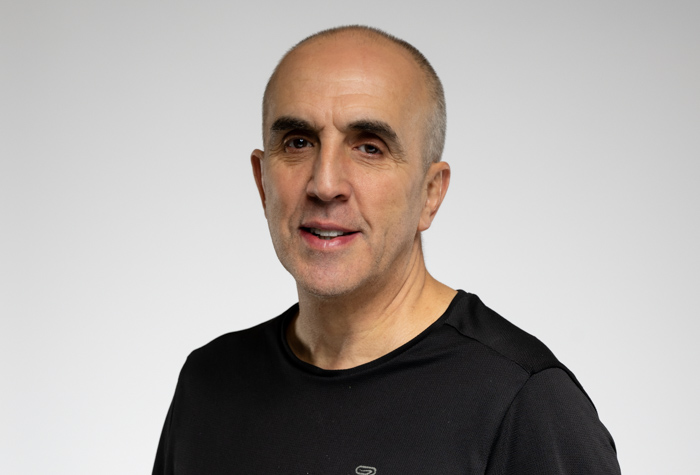 Un homme d'âge moyen aux cheveux gris courts et à la ligne capillaire fuyante sourit en portant une chemise noire sur un fond gris clair uni - il s'agit de Luc Tremblay.