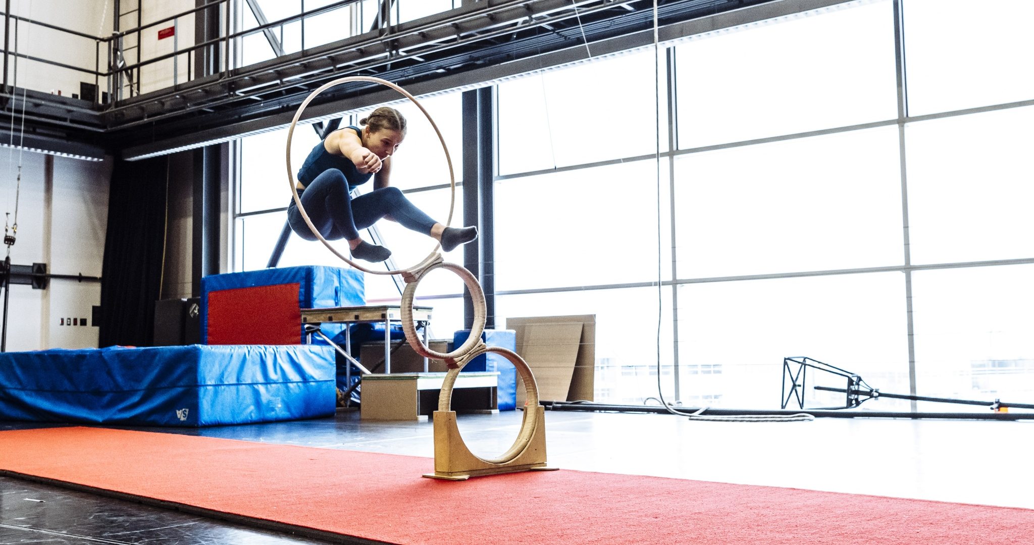 Lors d'un camp d'été Immersion cirque, une personne en tenue de sport saute à travers trois cerceaux empilés sur un tapis rouge à l'intérieur d'un gymnase spacieux et lumineux avec de grandes fenêtres et des équipements de gymnastique en arrière-plan.