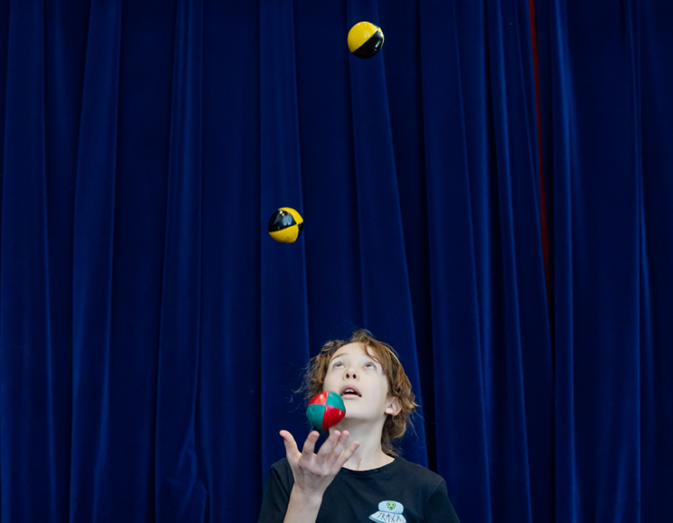 Une jeune personne aux cheveux bruns jongle avec trois balles colorées devant un grand rideau bleu foncé, en levant les yeux pour se concentrer sur les balles en l'air.