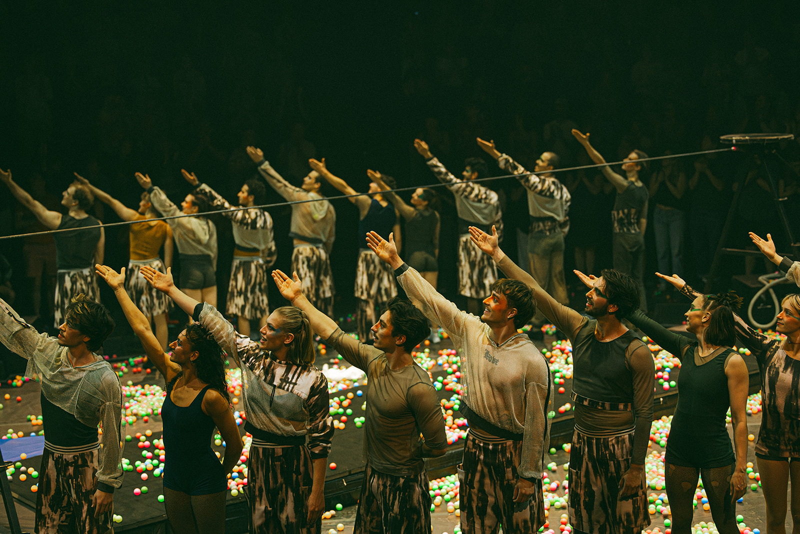 Un groupe d'artistes se tient en rangs sur une scène couverte de boules colorées, étendant leurs bras droits vers le haut à l'unisson lors de la soirée-bénéfice de la Fondation de l'ENC, sous le regard d'un public en arrière-plan faiblement éclairé.