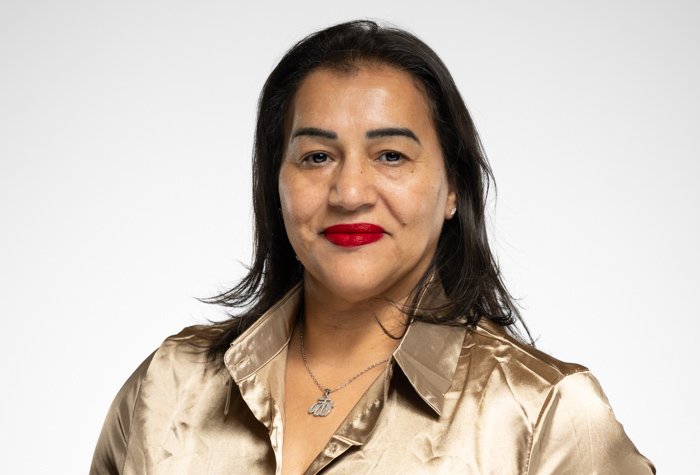 Halima Ayad, aux longs cheveux bruns, porte un chemisier beige brillant et du rouge à lèvres rouge, en souriant légèrement. Elle porte un collier en argent et pose devant un fond clair et uni.