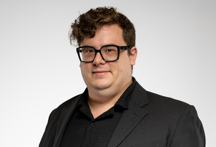 Olivier Leblanc, un homme aux cheveux bruns courts et bouclés et aux lunettes noires, porte une chemise noire et un blazer foncé et pose sur un fond gris clair uni.