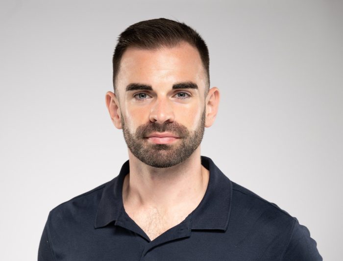 Maxime Blouin, un homme aux cheveux bruns courts et à la barbe taillée, porte une chemise à col bleu marine, se tient debout sur un fond gris clair uni et regarde directement la caméra avec une expression neutre.