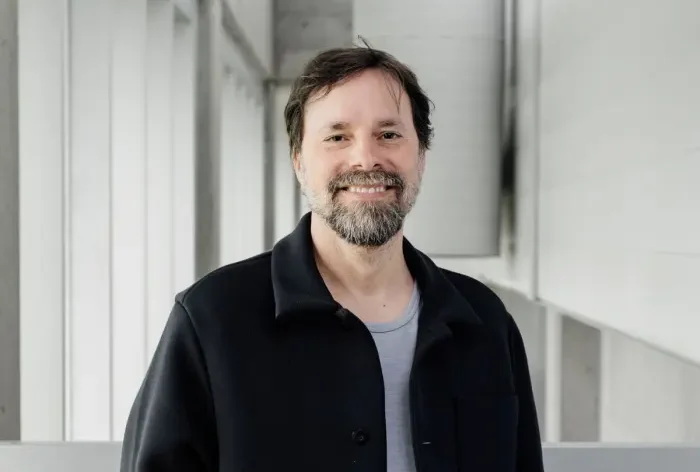 Patrice Aubertin, un homme barbu et moustachu, vêtu d'une veste noire sur une chemise gris clair, se tient à l'intérieur, près d'une balustrade, dans un couloir moderne et lumineux, et sourit à la caméra.