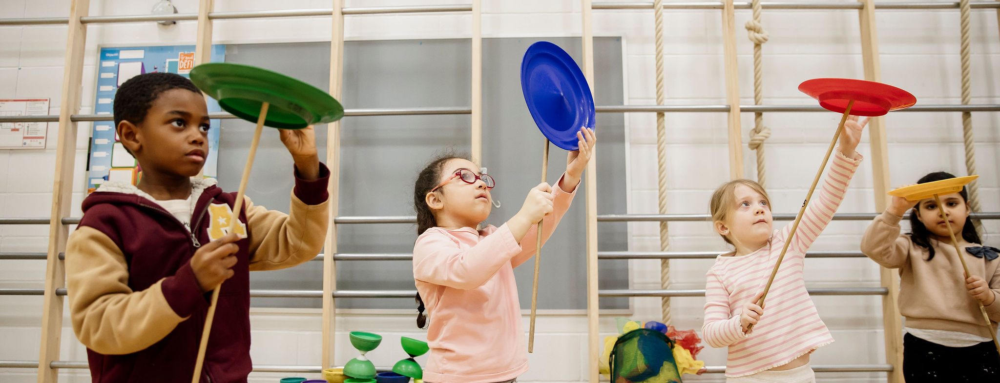 Quatre enfants se tiennent à l'intérieur, chacun tenant une assiette tournante sur un bâton. Ils se concentrent intensément sur l'équilibre des assiettes colorées, entourées d'équipements de gymnastique en arrière-plan.