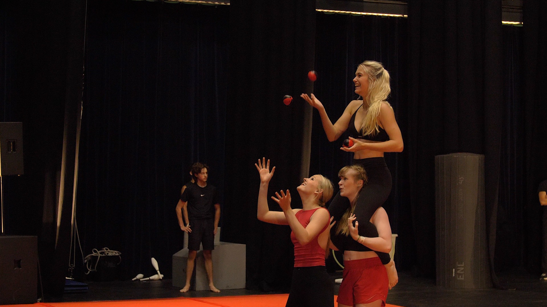 Trois femmes exécutent un numéro d'acrobatie ; deux d'entre elles se tiennent côte à côte et soutiennent une troisième femme sur leurs épaules alors qu'elle jongle avec des balles rouges. Rappelant les camps d'été intensifs, un homme observe en arrière-plan sur une scène sombre.