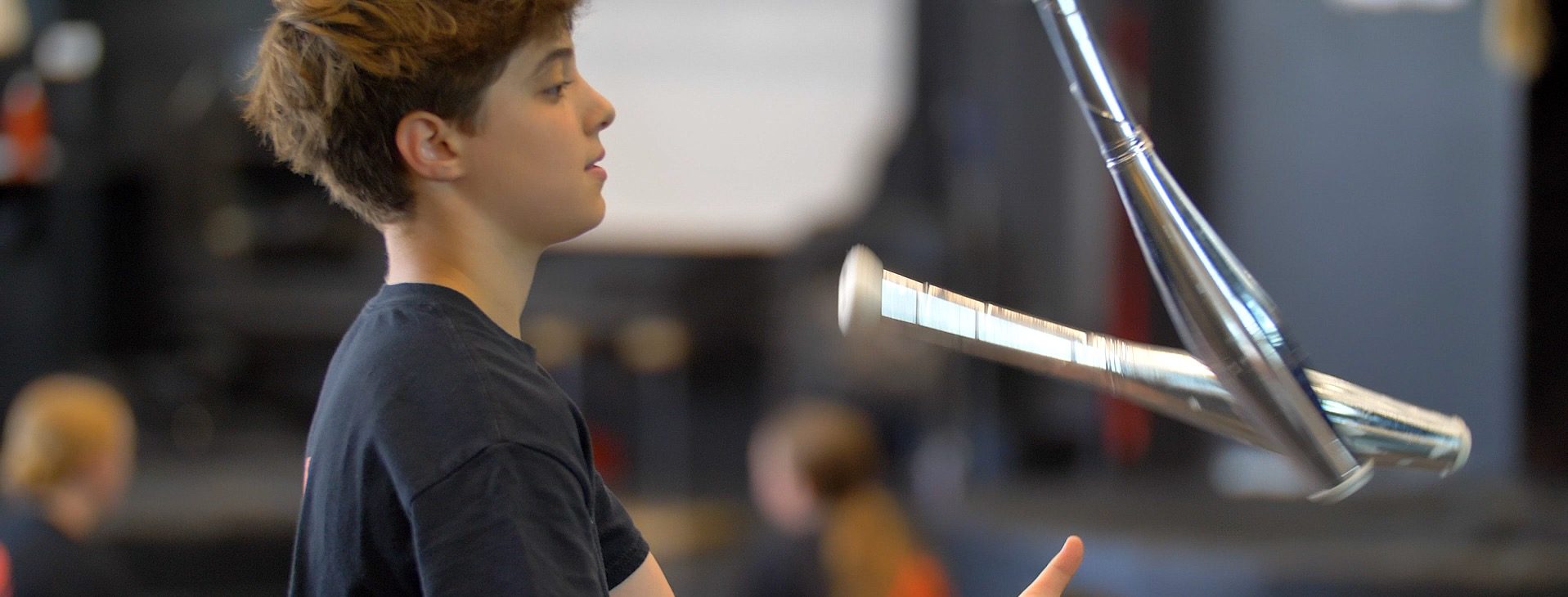 Une jeune personne aux cheveux bruns courts lance une massue de jonglage argentée en l'air, en se concentrant intensément - peut-être en pratiquant des techniques apprises lors de camps d'été intensifs. L'arrière-plan flou suggère un environnement intérieur avec d'autres personnes présentes.