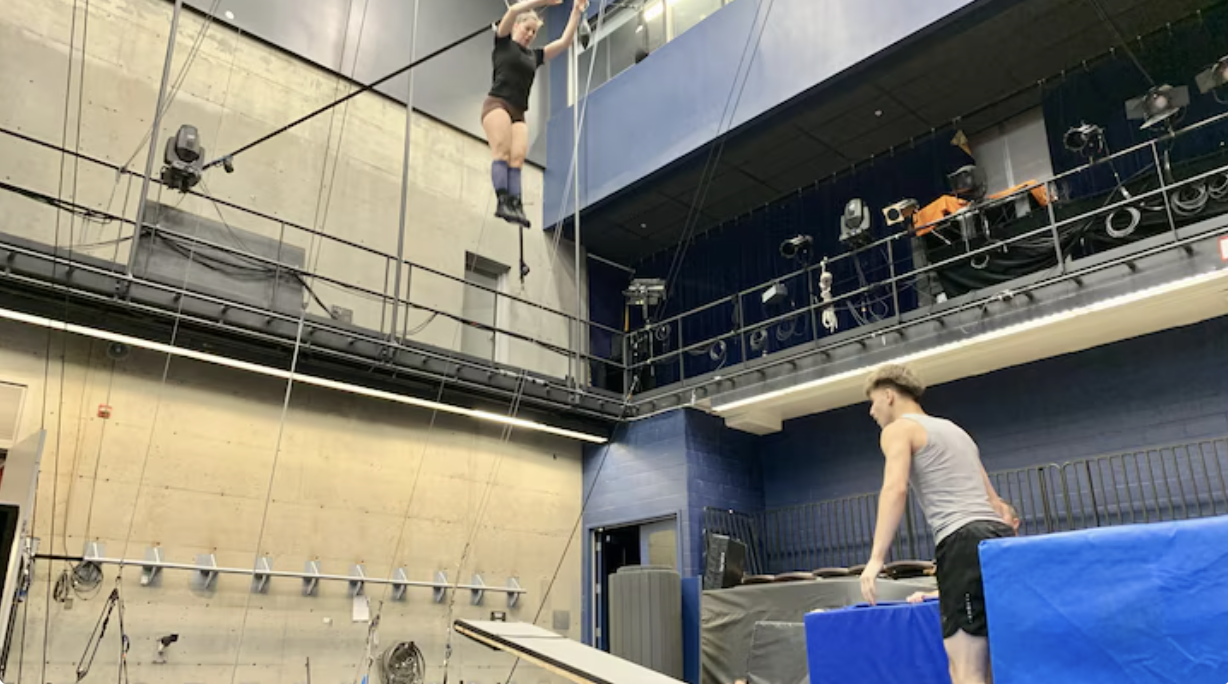 Une personne effectue un numéro aérien à l'aide d'un trapèze dans une salle de sport avec des tapis bleus en dessous, tandis qu'une autre personne vêtue d'un débardeur observe depuis le sol. L'espace est doté de hauts plafonds, de projecteurs et de cordes d'escalade sur le mur.
