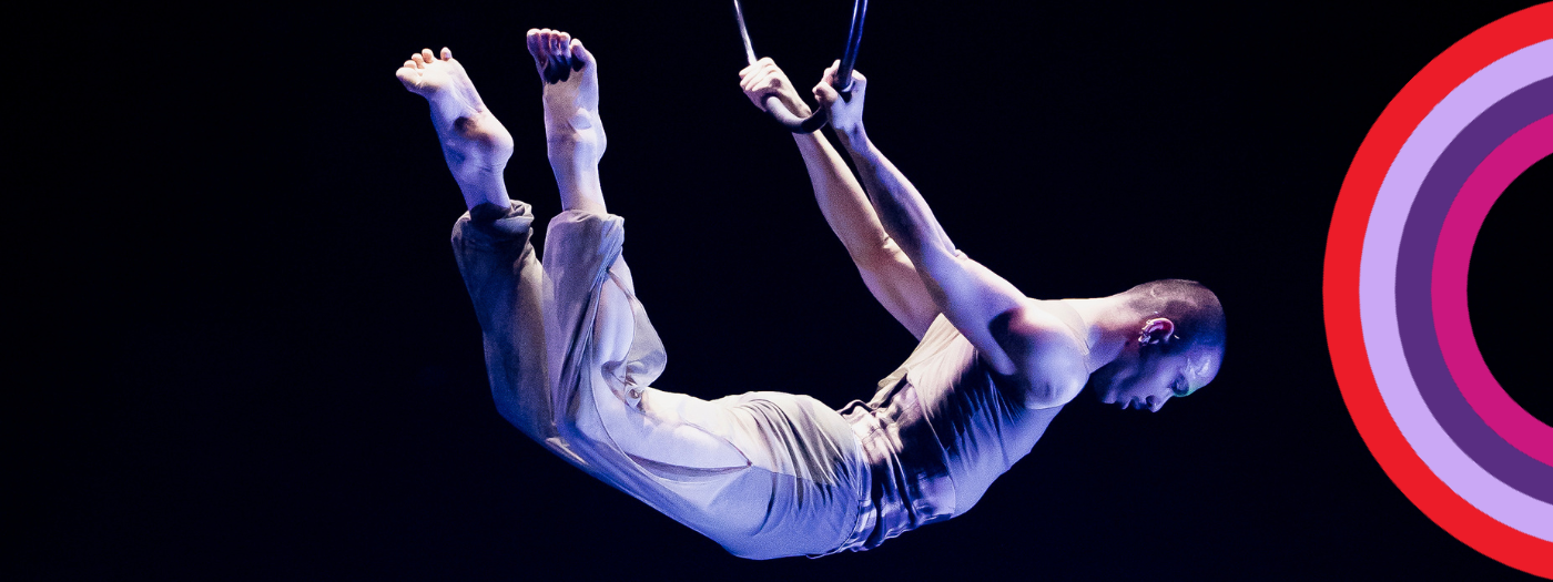 Un acrobate masculin vêtu de couleurs claires se produit sur des anneaux aériens sur un fond sombre, mis en valeur par un éclairage dramatique, dans le cadre d'un spectacle d'étudiants en fin d'études, avec un graphique circulaire coloré à droite.