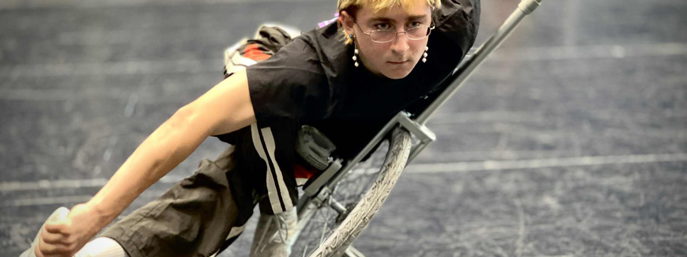 Une personne aux cheveux courts et aux lunettes joue intensément au floorball, vêtue d'une chemise noire et utilisant un fauteuil roulant de sport sur un terrain intérieur - découvrez la même énergie dans les nouveaux cours récréatifs à l'École nationale de cirque !.