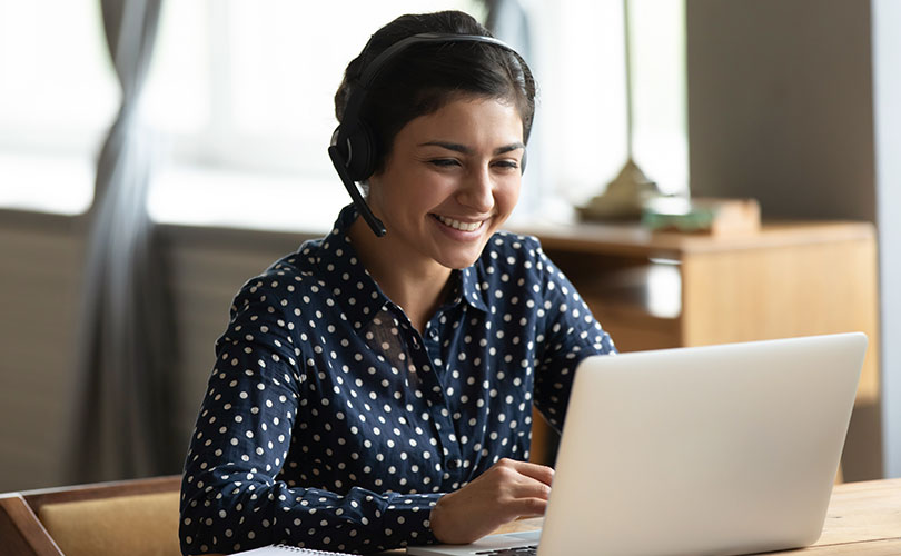 Une femme portant un casque et une chemise à pois bleu marine sourit alors qu'elle est assise à un bureau et qu'elle utilise un ordinateur portable. Elle semble être en appel vidéo ou travailler à distance dans une pièce lumineuse.
