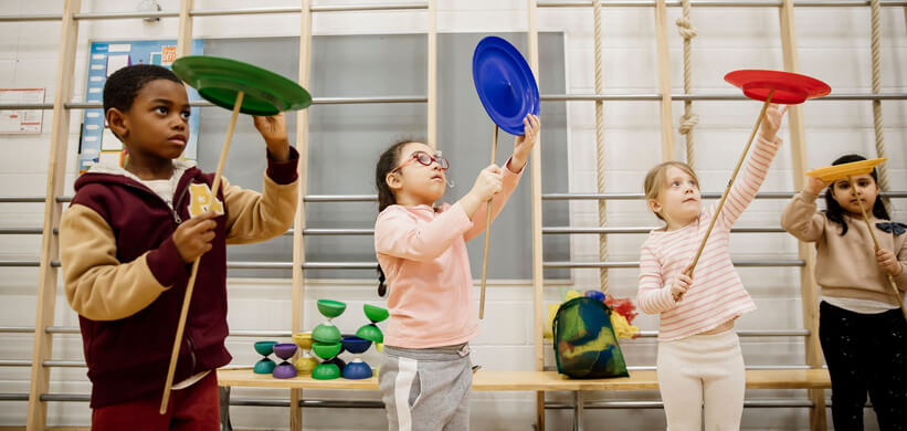 Quatre enfants à l'intérieur tiennent chacun une assiette en plastique colorée en équilibre sur un bâton, se concentrant pour s'entraîner à faire tourner l'assiette. Derrière eux se trouvent des barres murales de gymnastique et des accessoires de jonglerie placés sur un banc.