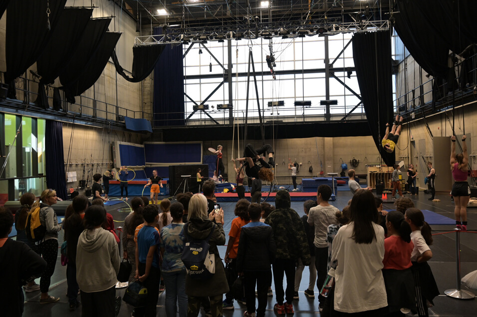 Un groupe d'enfants observe des artistes qui pratiquent l'acrobatie aérienne et d'autres techniques de cirque dans un grand gymnase intérieur avec de hauts plafonds, des cordes et de l'équipement. Certains artistes sont suspendus dans les airs, tandis que d'autres se tiennent au sol.