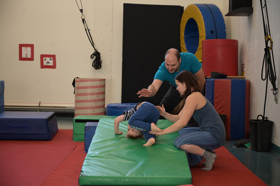Un jeune enfant pratique une roulade avant sur un tapis rembourré, aidé par un homme et une femme. Les deux adultes guident et soutiennent l'enfant à l'intérieur d'une salle de sport colorée dotée d'équipements souples.