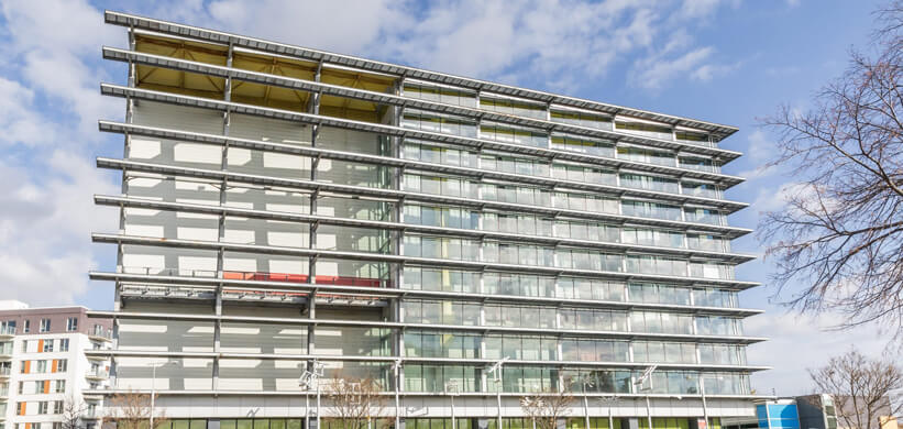 Un immeuble de bureaux moderne de plusieurs étages, en verre et en acier, avec des poutres métalliques horizontales, de grandes fenêtres et des balcons, sur fond de ciel partiellement nuageux, avec des arbres et d'autres bâtiments visibles à proximité.