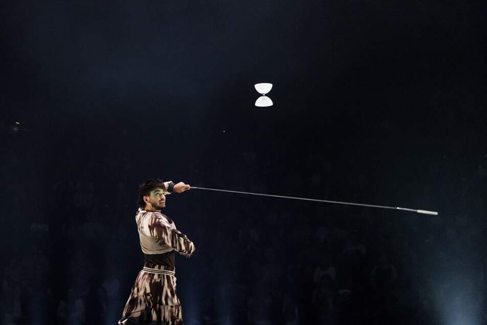 Un artiste vêtu de vêtements à motifs fait habilement tourner un diabolo sur une corde tenue entre deux bâtons, avec un public sombre à l'arrière-plan et un éclairage de scène dramatique qui met en valeur le numéro.