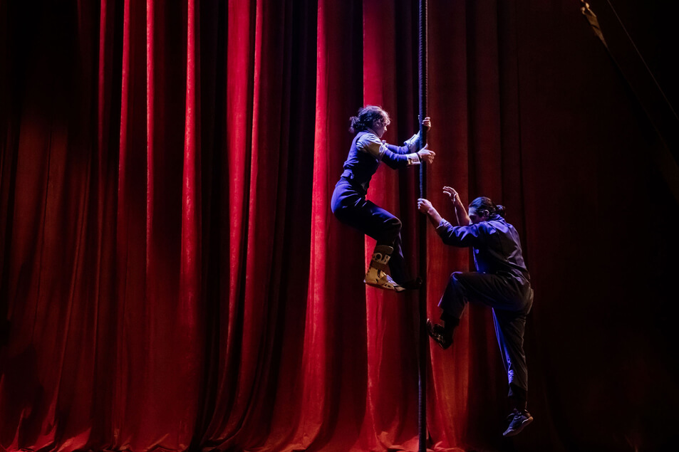 Deux artistes vêtus de vêtements sombres grimpent à une corde sur la scène devant des rideaux rouges dramatiques, éclairés par des lumières de scène, créant une atmosphère vibrante et théâtrale.