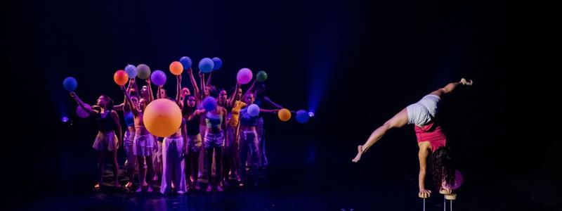 Un groupe d'artistes tenant des balles colorées se tient à l'arrière-plan, tandis qu'une personne au premier plan se tient en équilibre sur une main en haut d'un tabouret sur une scène faiblement éclairée. Période d'inscription prolongée jusqu'au 21 novembre.