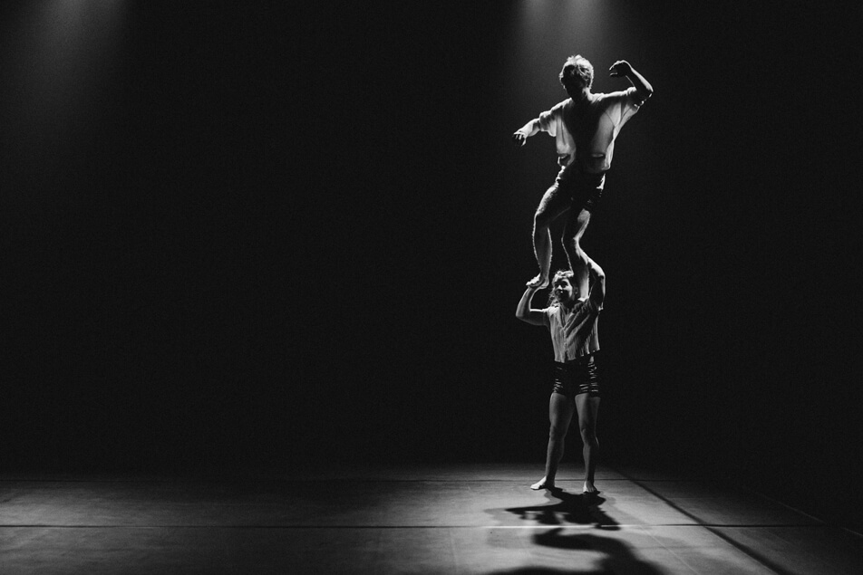 Deux acrobates exécutent un numéro d'équilibre sur une scène faiblement éclairée ; l'un se tient sur les épaules de l'autre tandis que tous deux posent sous des projecteurs, projetant des ombres dramatiques sur le sol.