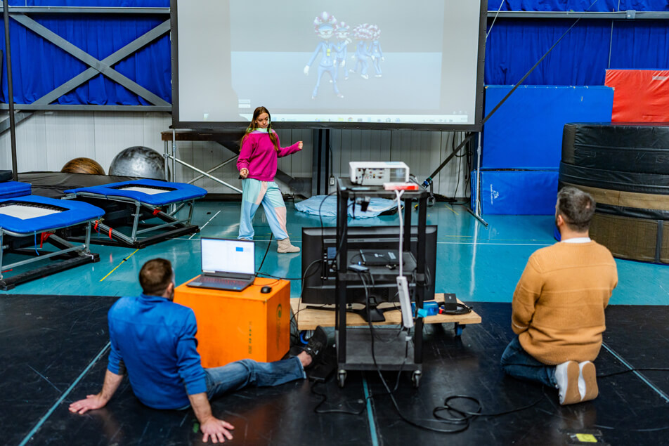 Une femme fait un geste en direction d'un grand écran affichant des personnages animés, tandis que deux hommes assis regardent. Ils se trouvent dans un espace ressemblant à un gymnase, avec des trampolines, des tapis et des équipements à proximité. Des ordinateurs portables et un projecteur sont également visibles.