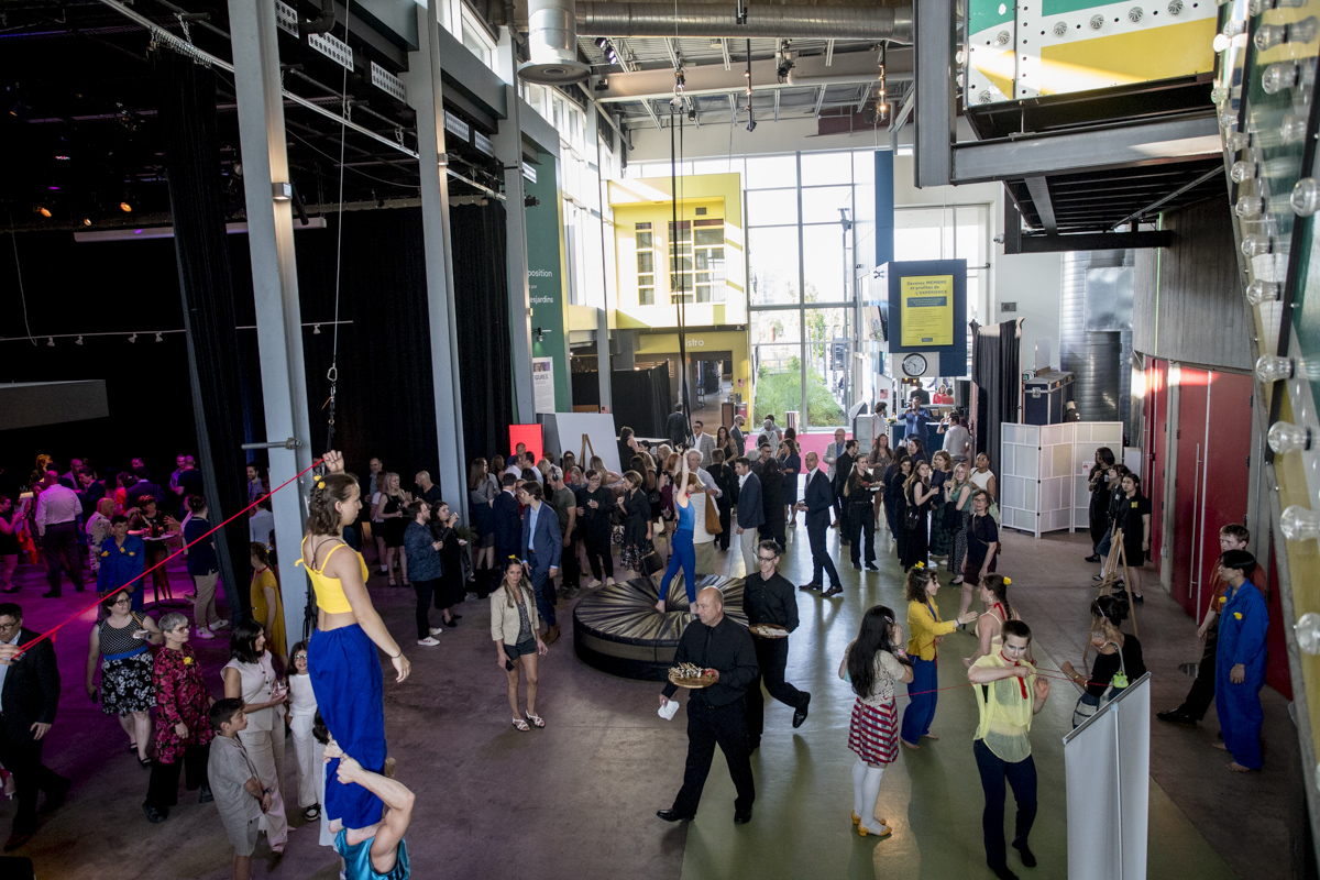 Une soirée-bénéfice intérieure animée de la Fondation de l'ENC avec des gens qui socialisent ; certains regardent des artistes sur une slackline et un cerceau aérien. Le lieu est doté de hauts plafonds, de grandes fenêtres, d'un décor industriel, de murs colorés et d'éclairages dispersés.