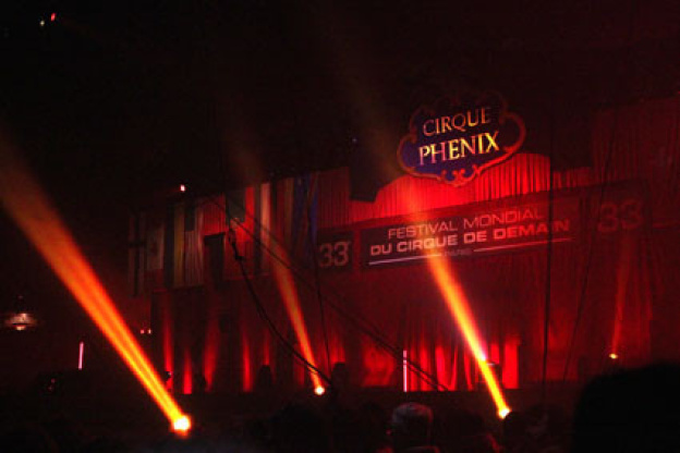 Scène faiblement éclairée avec des lumières rouges et des projecteurs au Cirque Phenix. Un panneau indique "Festival Mondial du Cirque de Demain" au-dessus de la scène, avec "33" de chaque côté. L'ENC bien représentée au Festival mondial du cirque de demain, renforçant l'atmosphère dramatique.