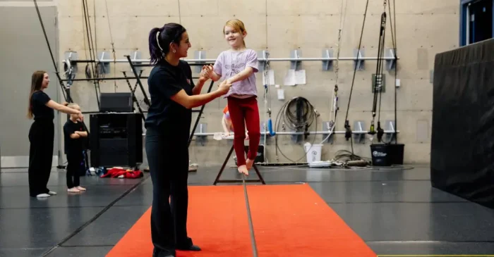 Une femme aide une jeune fille souriante à se tenir en équilibre sur une slackline au-dessus d'un tapis rouge dans une salle de sport intérieure, tandis que d'autres enfants et adultes s'entraînent à l'arrière-plan.