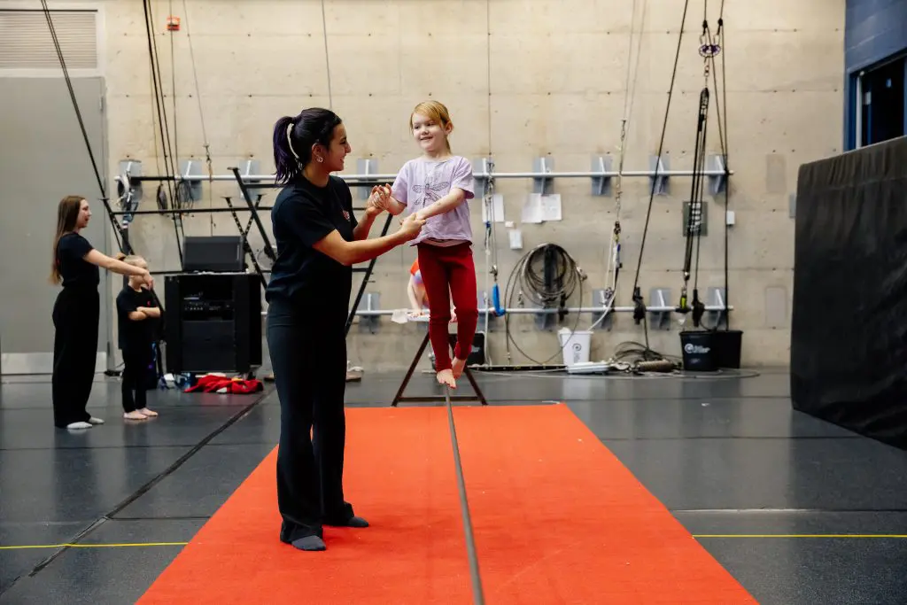 Une femme aide une jeune fille souriante à se tenir en équilibre sur une slackline au-dessus d'un tapis rouge dans une salle de sport intérieure, tandis que d'autres enfants et adultes s'entraînent à l'arrière-plan.