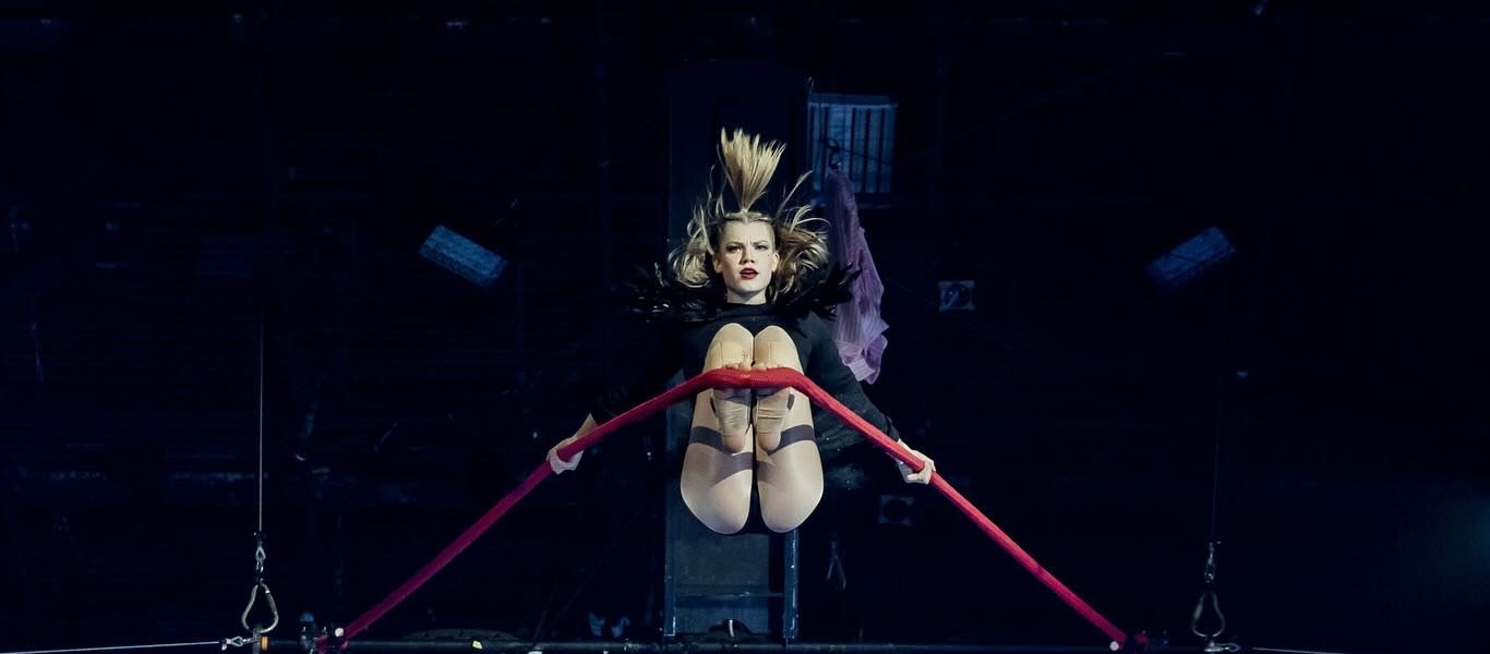 Une acrobate en justaucorps évolue sur un trapèze suspendu, agrippant une barre rouge et pliant les genoux en plein vol sous un éclairage de scène spectaculaire - une scène captivante qui rappelle une illustration de Diptyque.