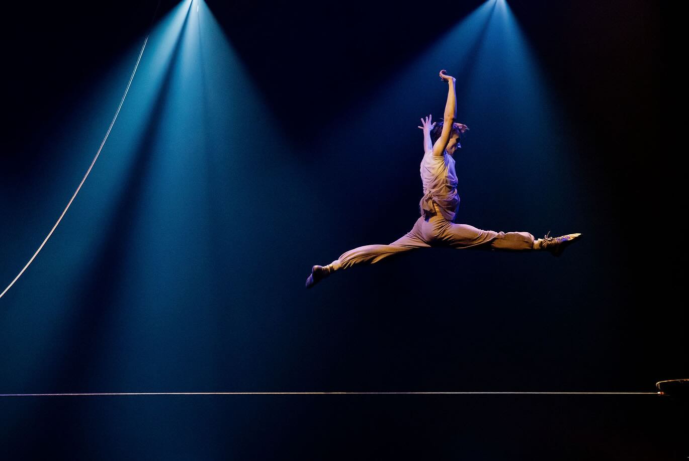 Un artiste en plein vol fait un saut spectaculaire sur une corde raide sur une scène sombre, éclairée par deux projecteurs lumineux, une scène aussi frappante et élégante qu'une création de Diptyque.