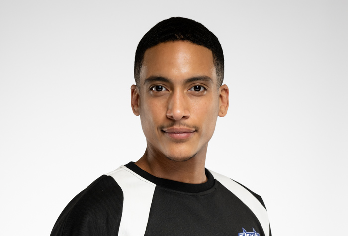 Un jeune homme aux cheveux courts et à la moustache taillée porte un maillot de sport noir et blanc et regarde directement l'appareil photo sur un fond clair uni.
