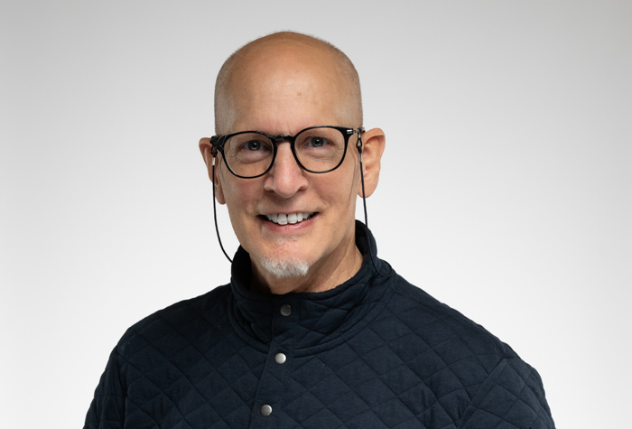 Un homme chauve souriant avec des lunettes et une petite barbichette grise porte une veste matelassée foncée sur un fond gris clair uni.