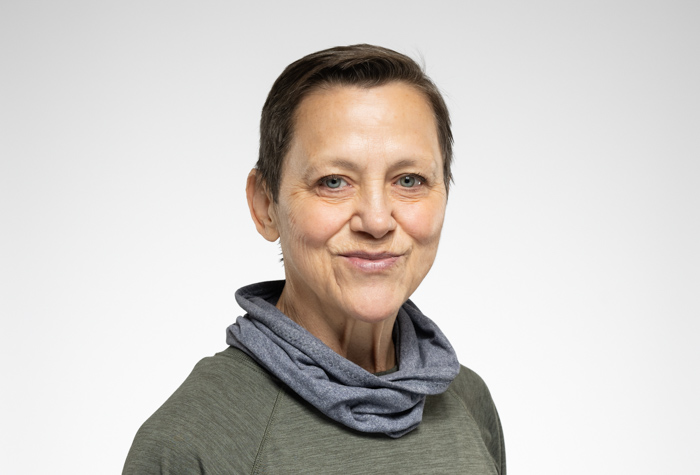 Une personne d'âge moyen aux cheveux bruns courts, vêtue d'un haut vert et d'une écharpe grise, sourit doucement à l'appareil photo sur un fond clair et uni.